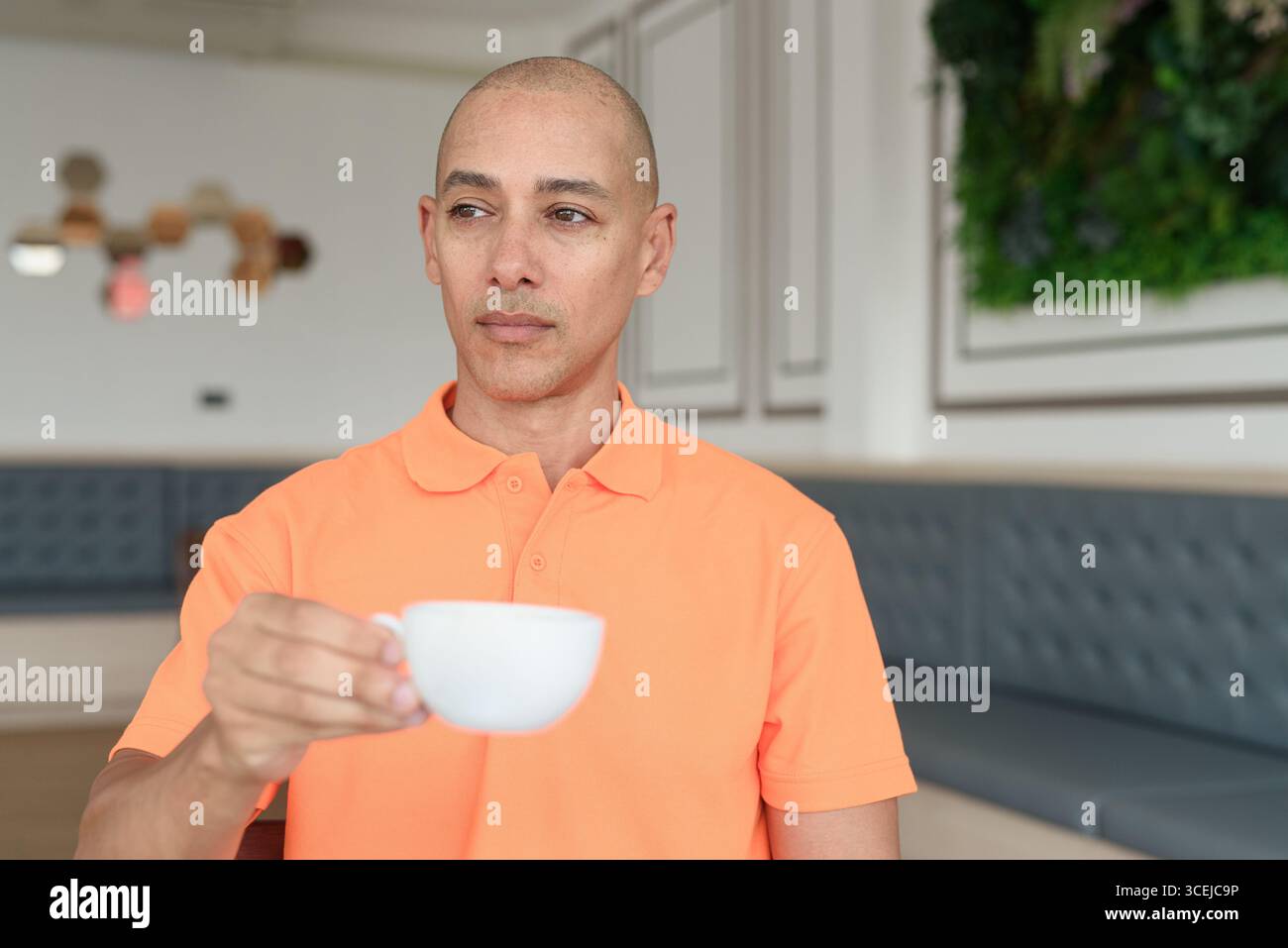 Bel homme chauve italien d'âge moyen en polo orange assis à la table de café tenant une tasse de café. Concept de style de vie décontracté. Banque D'Images
