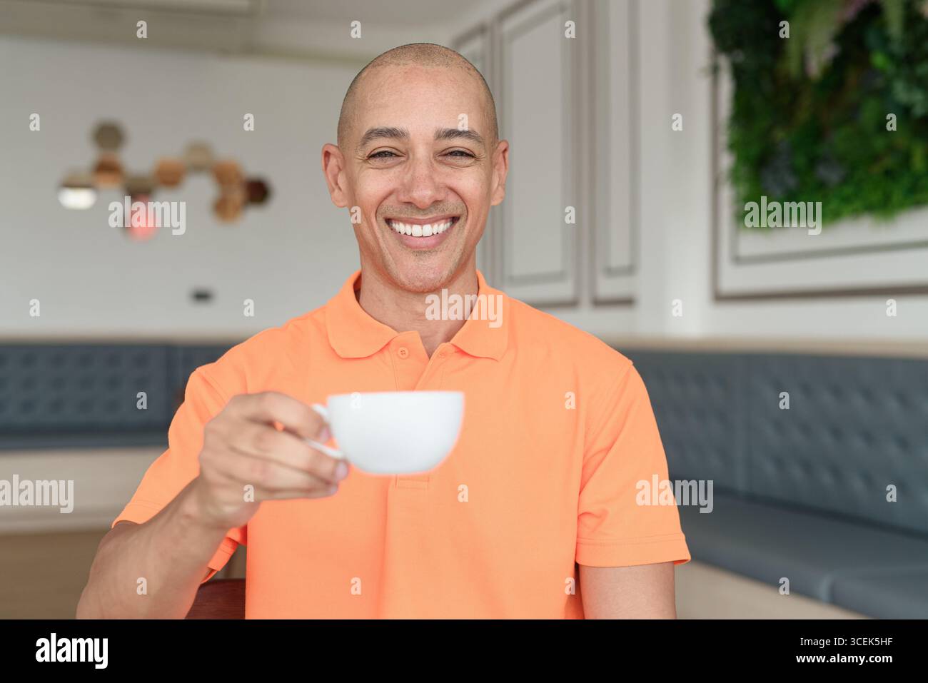 Bel homme chauve italien d'âge moyen souriant dans un polo orange assis à la table de café tenant une tasse de café. Concept de style de vie décontracté. Banque D'Images