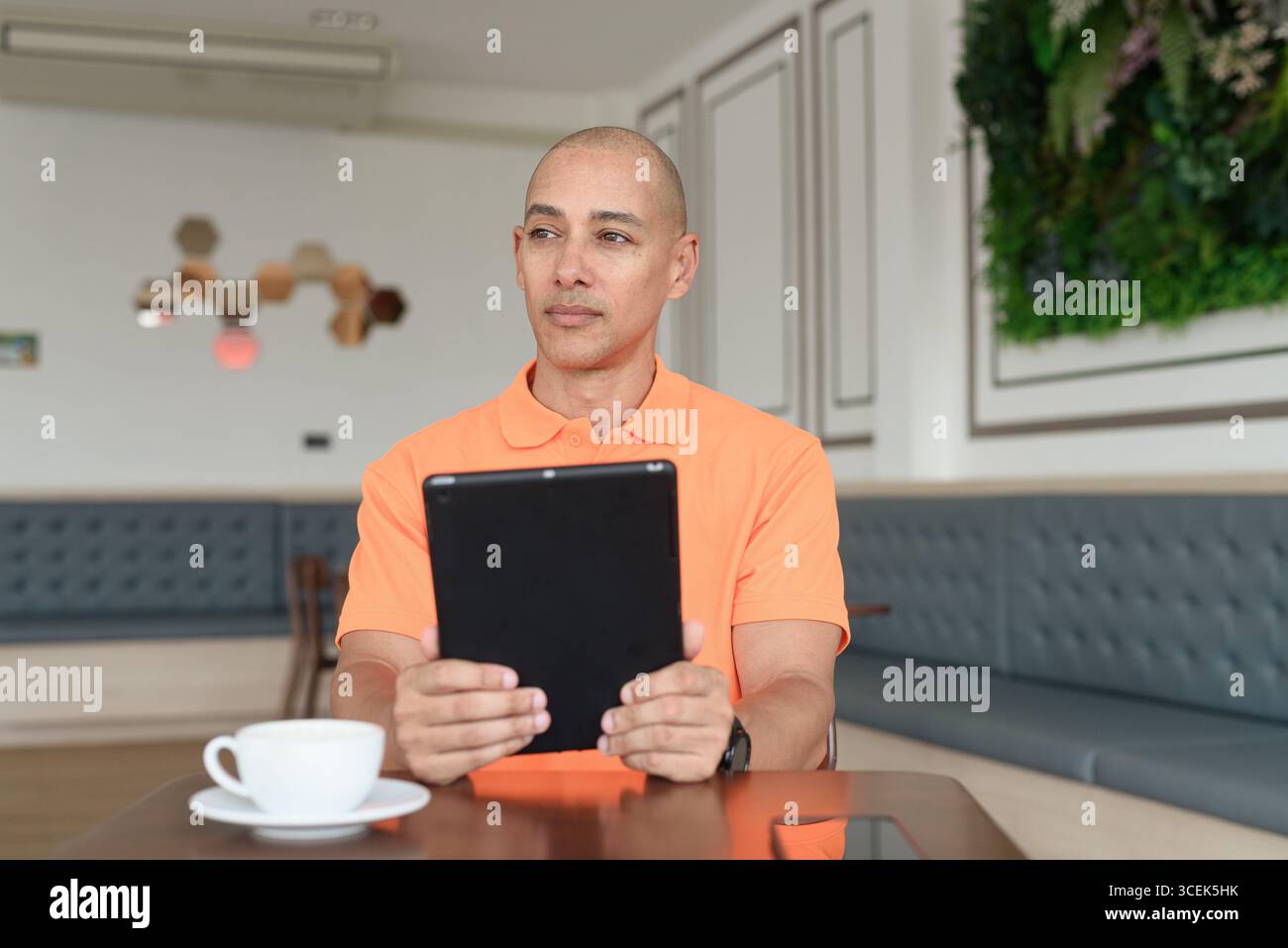 Bel homme d'âge moyen dans un polo orange à la table de café avec tasse à café, portant des écouteurs, et à l'aide d'un ordinateur tablette numérique. Technologie et cas Banque D'Images