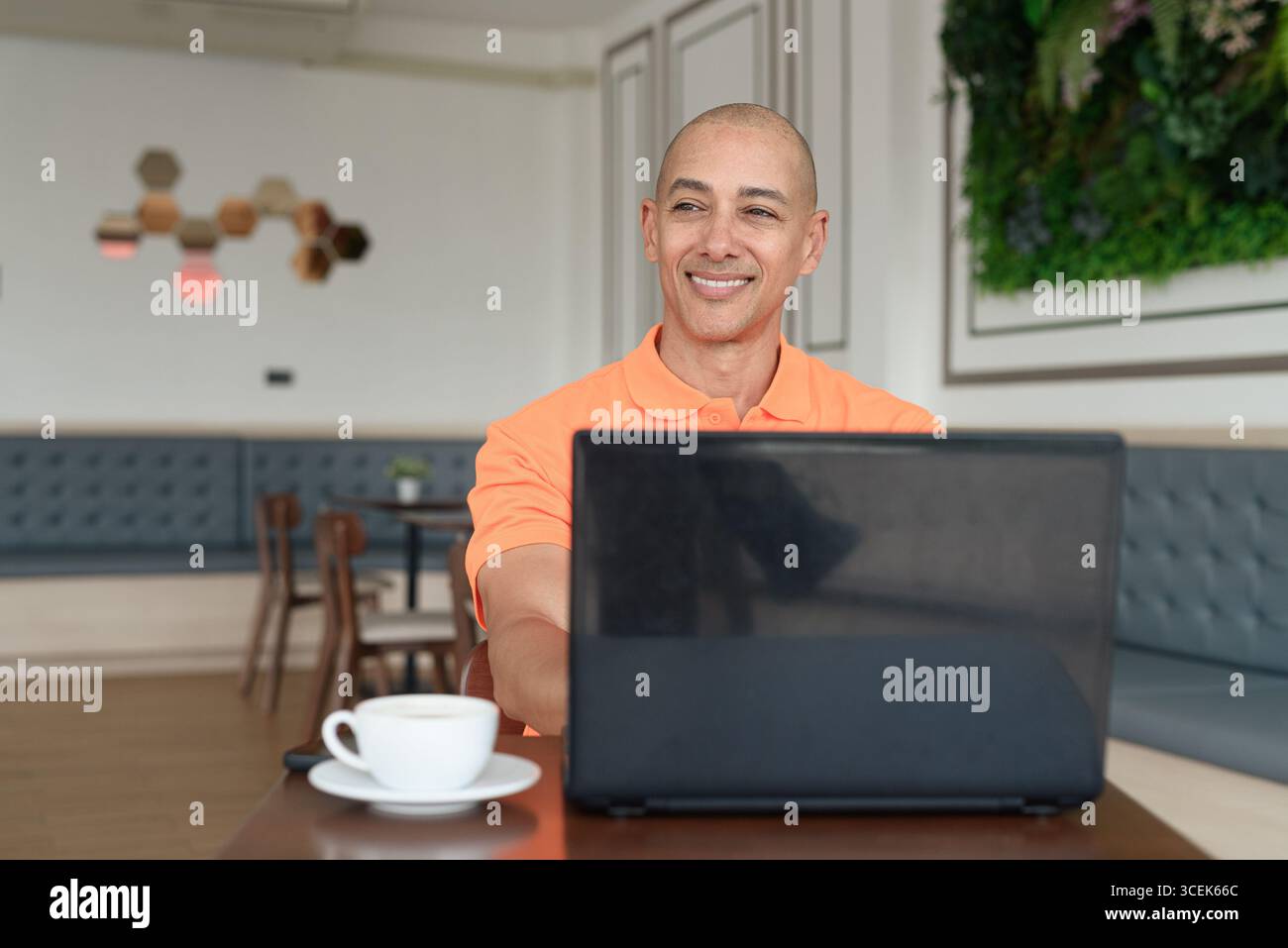 Bel homme hispanique chauve d'âge moyen en polo orange à la table de café avec tasse à café travaillant sur ordinateur portable à l'intérieur. Télétravail et technolo Banque D'Images