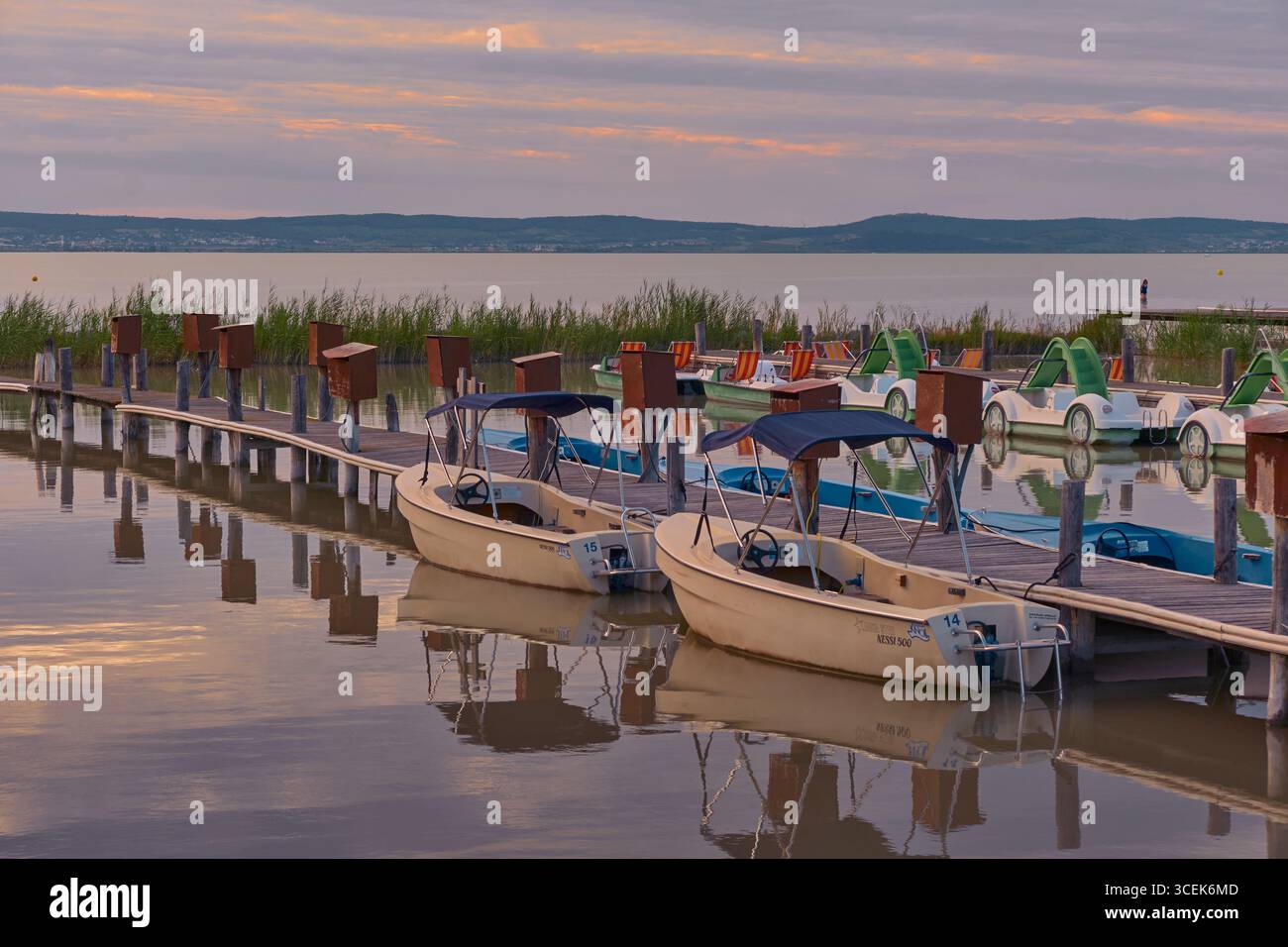 Quai avec de petits bateaux électriques amarrés dans le lac de Neusiedl, Autriche, entouré de verdure, sous un ciel de coucher de soleil pastel doux avec des tons roses et bleus. Banque D'Images