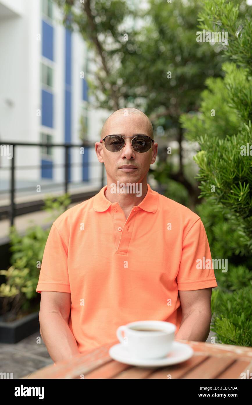 Beau visage d'homme chauve italien d'âge moyen dans un polo orange assis à la table du café à l'extérieur portant des lunettes de soleil. Concept de style de vie décontracté. Banque D'Images