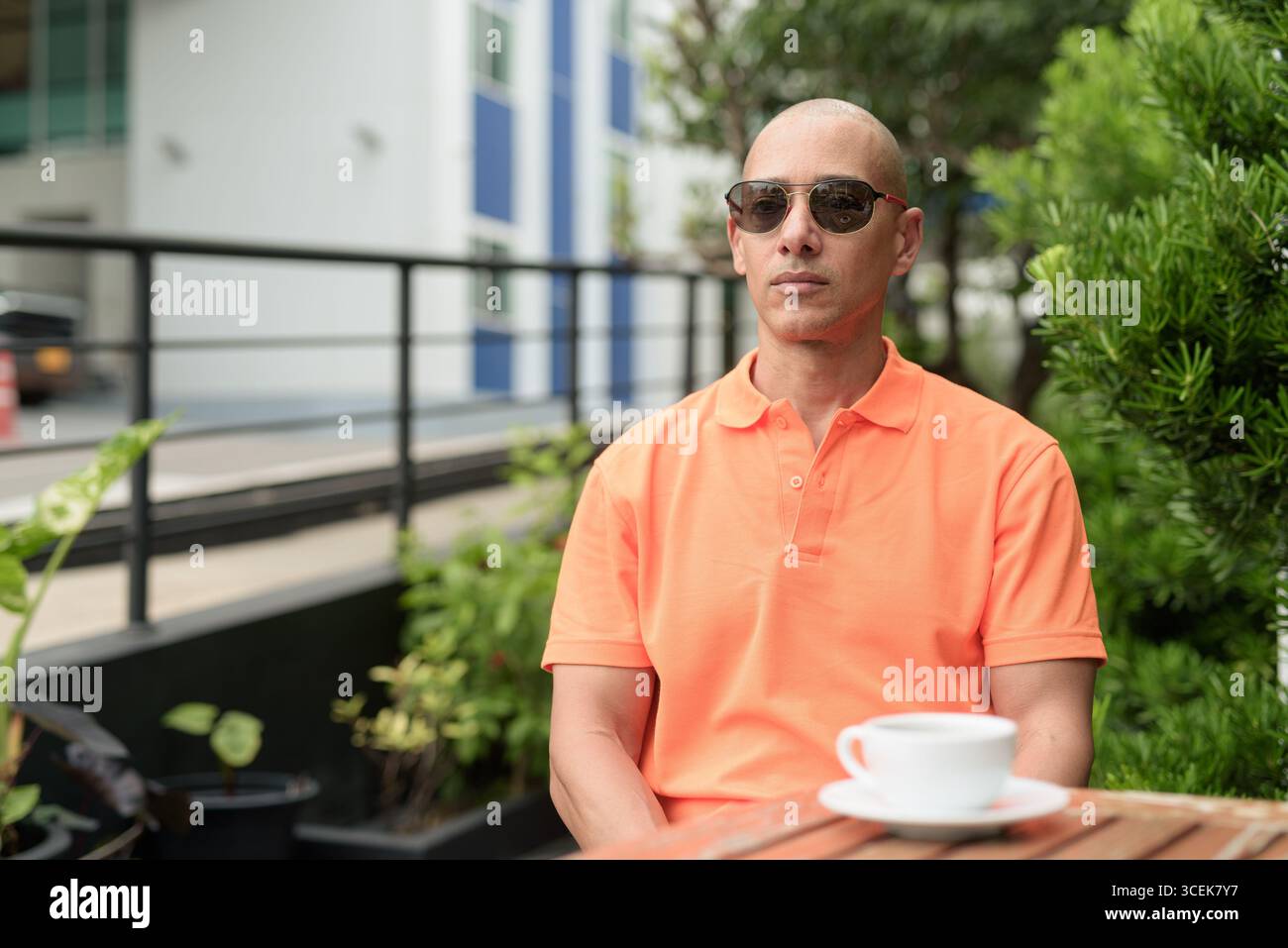 Beau visage d'homme chauve italien d'âge moyen dans un polo orange assis à la table du café à l'extérieur portant des lunettes de soleil. Concept de style de vie décontracté. Banque D'Images