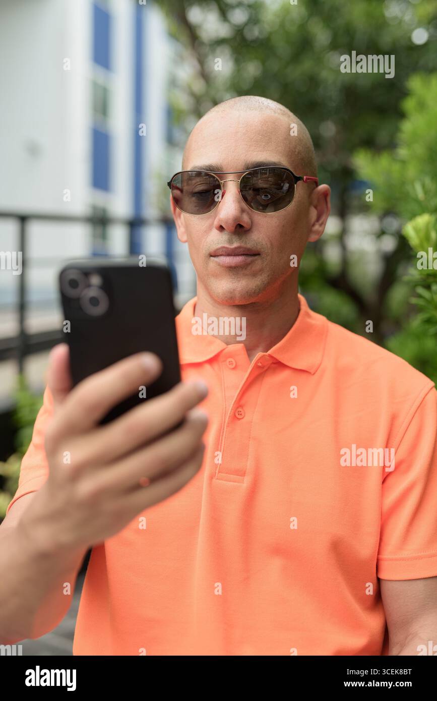 Beau visage d'homme chauve italien d'âge moyen dans un polo orange assis à la table du café à l'extérieur portant des lunettes de soleil et utilisant un téléphone portable. Vie décontractée Banque D'Images