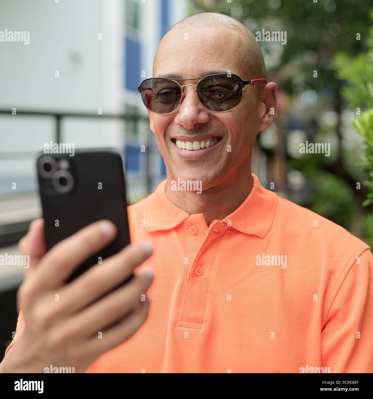 Beau visage d'homme chauve italien d'âge moyen dans un polo orange assis à la table du café à l'extérieur portant des lunettes de soleil et utilisant un téléphone portable. Vie décontractée Banque D'Images