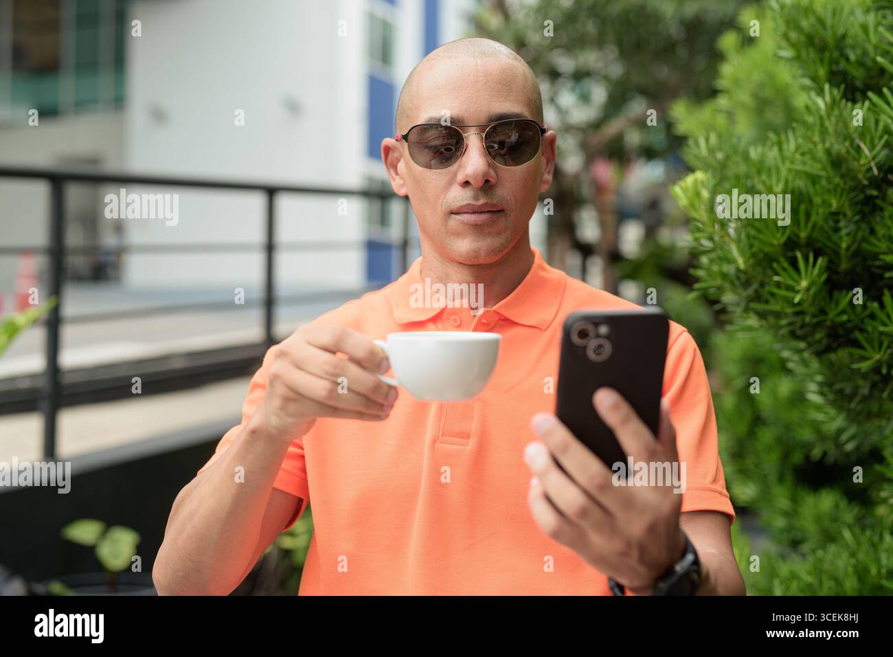 Beau visage d'homme chauve italien d'âge moyen dans un polo orange assis à la table du café à l'extérieur portant des lunettes de soleil et utilisant un téléphone portable. Vie décontractée Banque D'Images