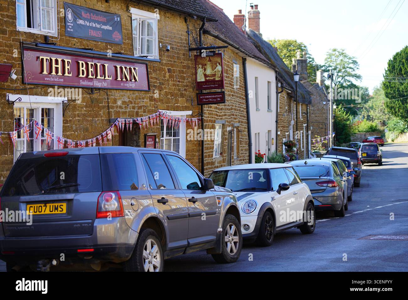 Pittoresque Village Street avec pub traditionnel et voitures garées un jour ensoleillé. Adderbury, Angleterre Banque D'Images