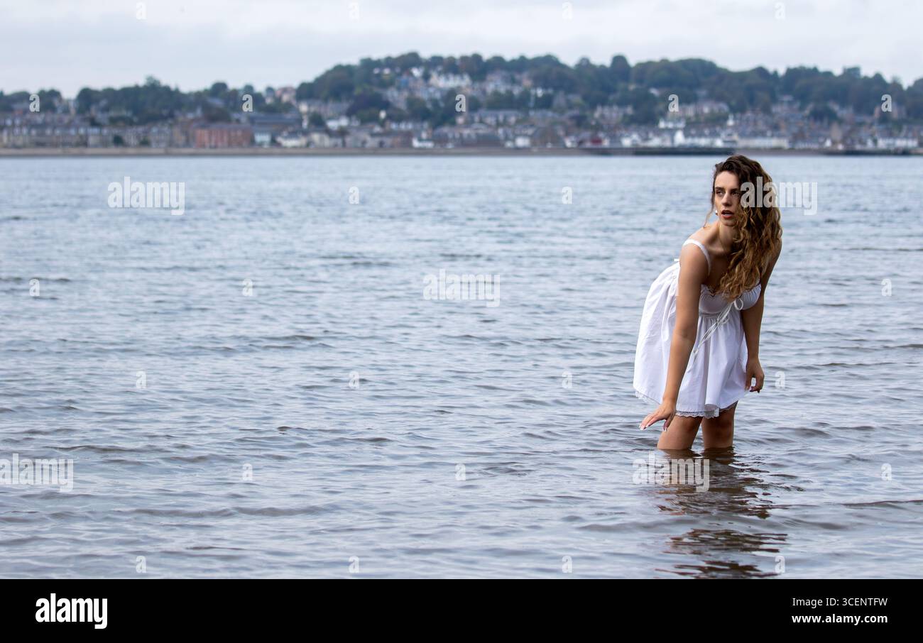 Tayport à Fife, en Écosse. Femme à la mode portant une robe blanche courte se tient dans la rivière Tay lors d'une séance photo sur le thème de la fantaisie l'après-midi, Royaume-Uni Banque D'Images