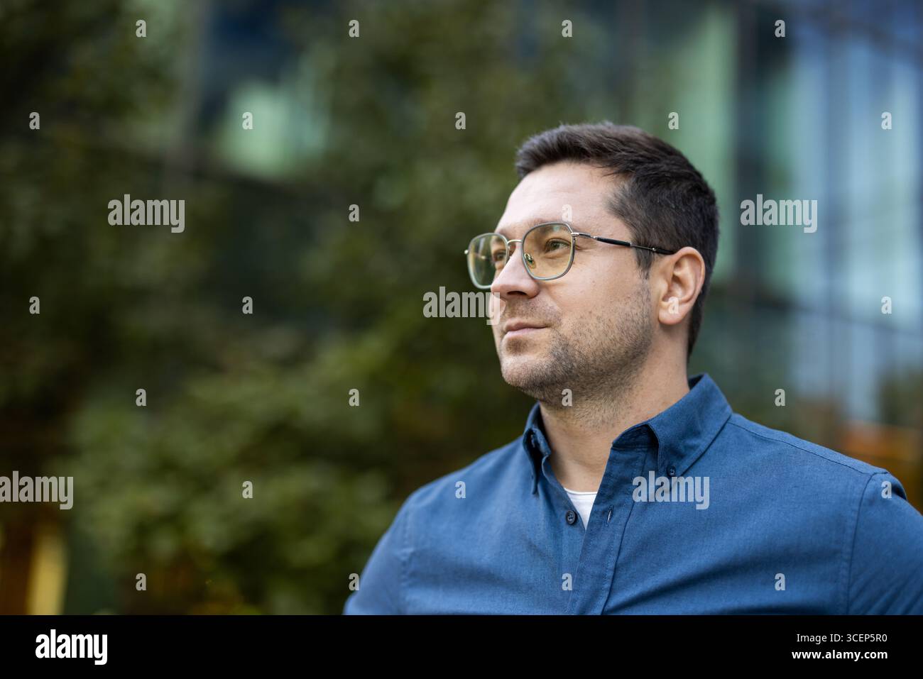 Un jeune professionnel confiant portant des lunettes debout à l'extérieur près d'un bâtiment moderne. Les environs comprennent des arbres et un extérieur en verre, créant une atmosphère professionnelle mais décontractée. Banque D'Images