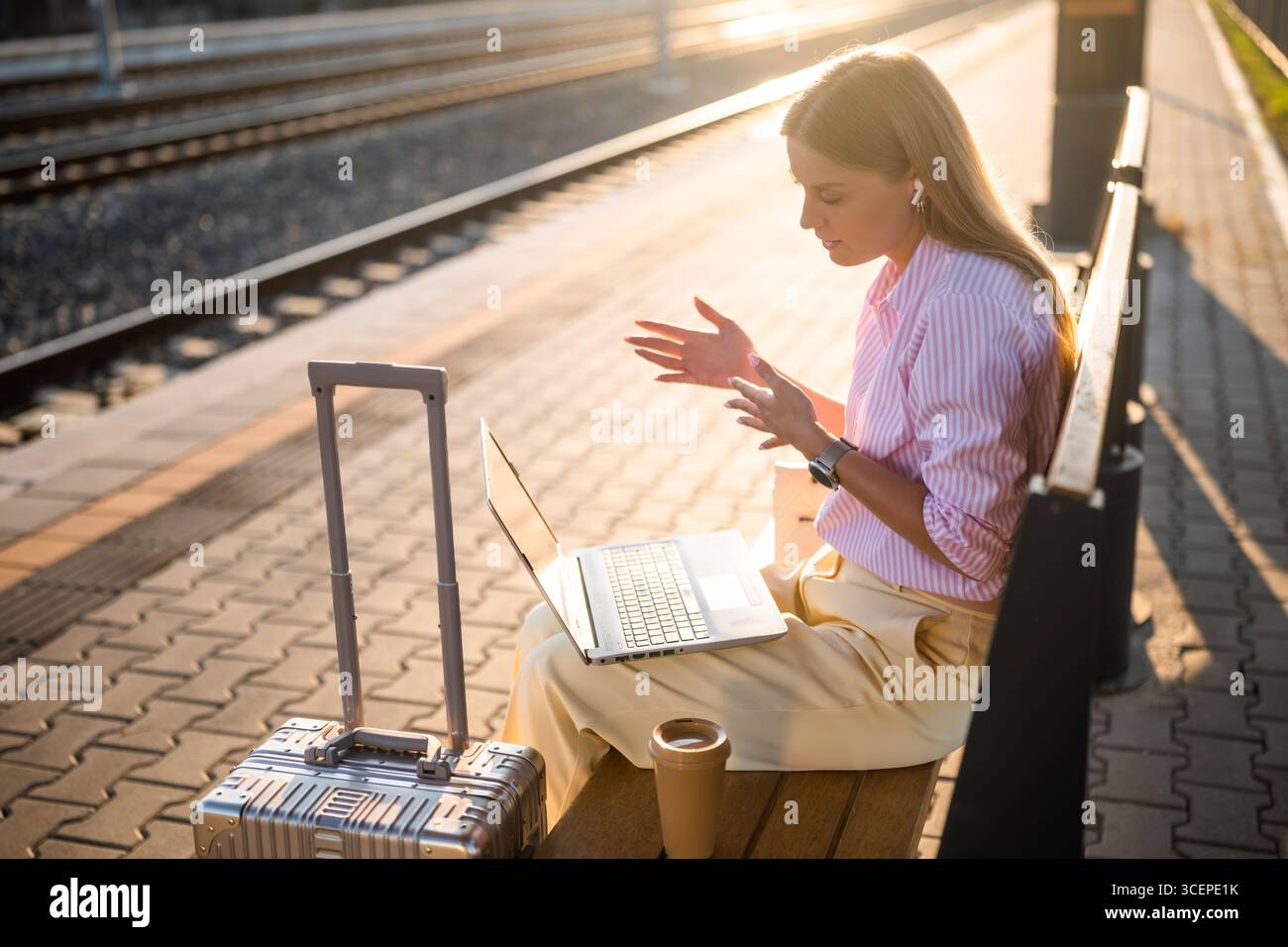 Femme d'affaires élégante utilisant un ordinateur portable et des écouteurs tout en étant assis sur un banc avec valise à la gare. Elle parle pendant un appel vidéo. Banque D'Images
