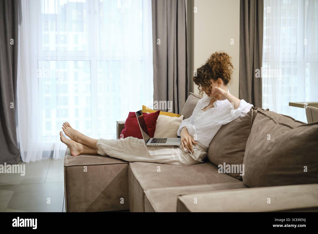 Une jeune femme aux cheveux bouclés se prélasse sur un canapé élégant dans un salon lumineux et regarde un film sur un ordinateur portable. L'atmosphère allie confort wi Banque D'Images