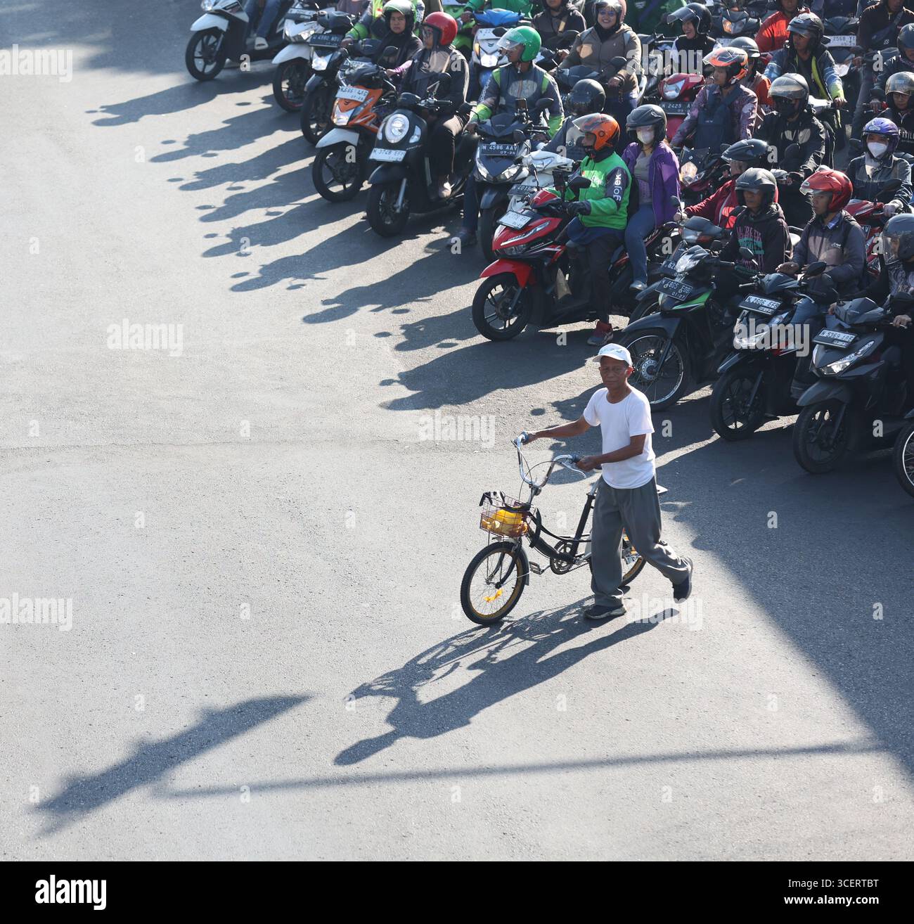 Un homme âgé passe son vélo devant des motocyclistes qui attendaient à un feu de circulation à Malang, Java oriental, Indonésie, le 1er août 2025. Banque D'Images