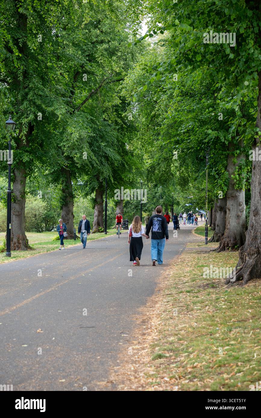 Un couple marche main dans la main le long d'un chemin bordé d'arbres dans un parc, entouré de verdure luxuriante et d'autres personnes appréciant la journée. Banque D'Images