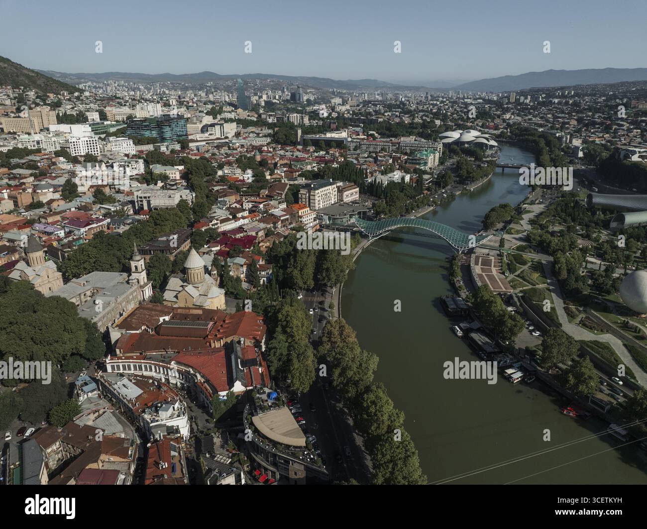 Vue aérienne de la rivière Kura serpentant à travers la ville, avec le Pont de la paix voûtant gracieusement au-dessus de l'eau, Tbilissi, Tbilissi, Géorgie. Banque D'Images
