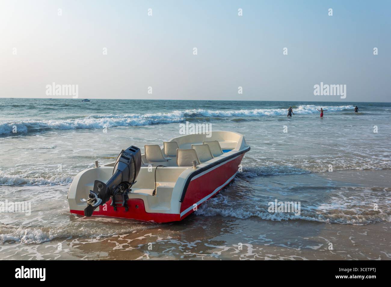 Bateau à moteur rouge et blanc garé sur une plage de sable avec des vagues douces et des gens nageant en arrière-plan, sous un ciel dégagé. Banque D'Images