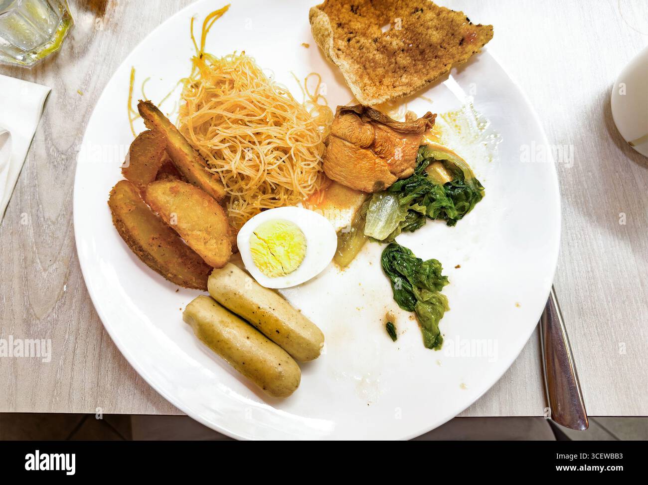Vue de dessus assiette d'aliments de petit déjeuner assortis, y compris les œufs, nouilles, quartiers de pommes de terre, légumes verts, saucisses, et du pain sur une table en bois Malacca malaisie Banque D'Images