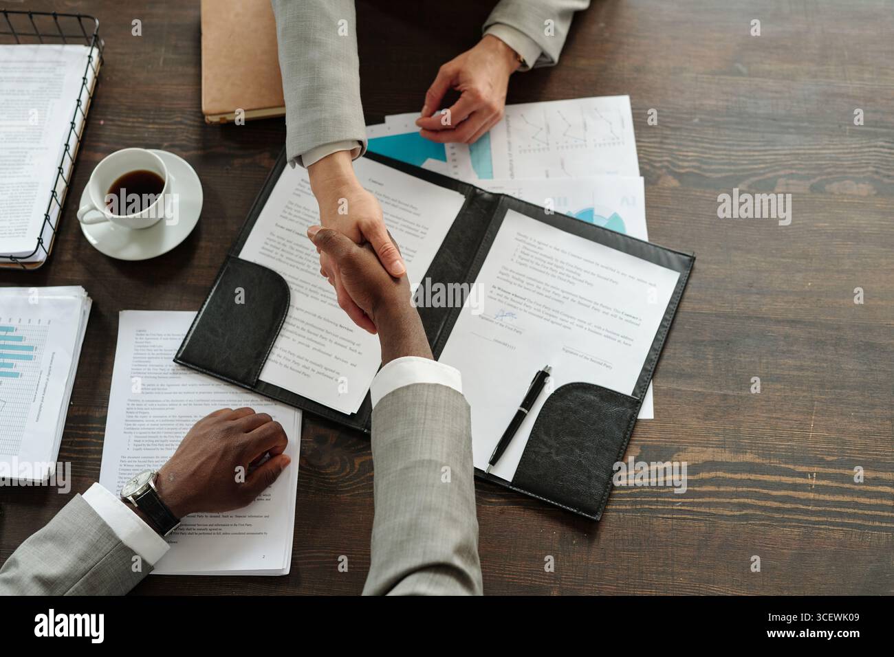 Femme caucasienne et homme noir serrant la main sur les documents contractuels sur le bureau en bois, les deux portant des vêtements d'affaires, tasse à café et graphiques financiers visibles, composition de vue de dessus Banque D'Images