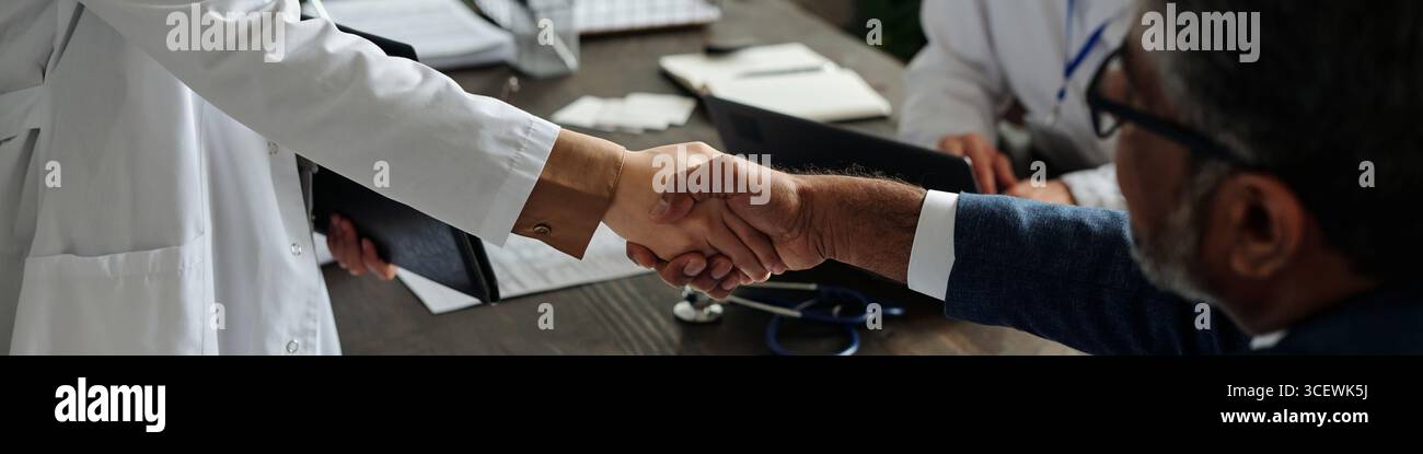 Homme caucasien d'âge moyen serrant la main avec femme caucasienne d'âge moyen médecin au bureau, professionnel de la santé tenant une presse-papiers, stéthoscope et ordinateur portable visible sur la table en bois Banque D'Images