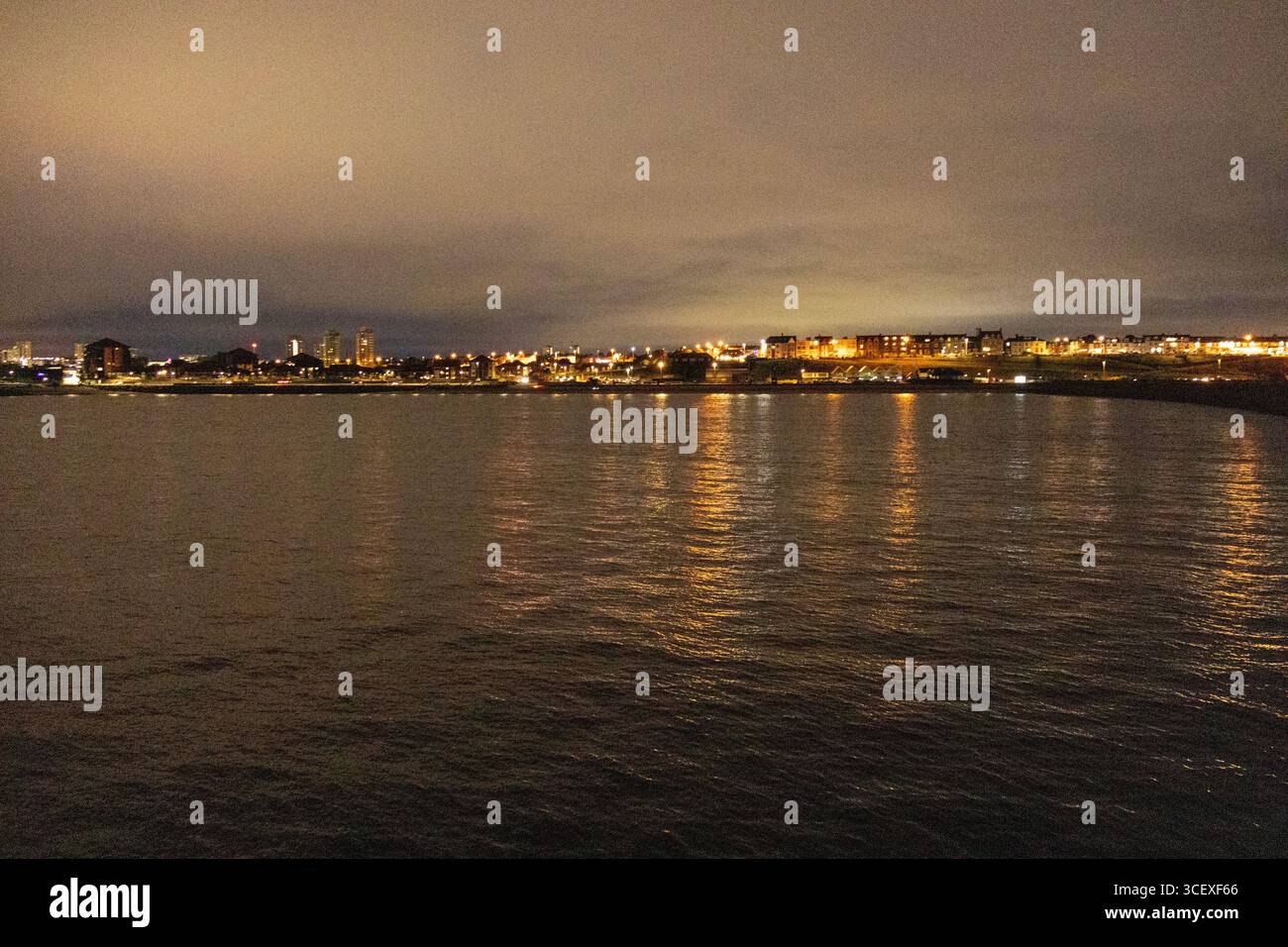 Vue nocturne des lumières de la ville reflétant une étendue d'eau calme, créant une ligne d'horizon urbaine lumineuse sous un ciel nuageux Banque D'Images