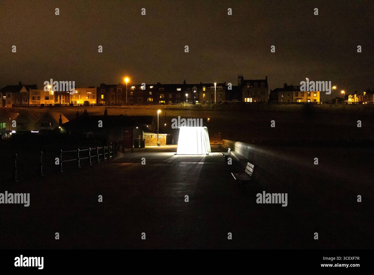 Une installation lumineuse incandescente la nuit sur une passerelle au bord de la rivière, avec des bancs, des lampadaires et des bâtiments résidentiels en arrière-plan. Pho de nuit urbaine Banque D'Images