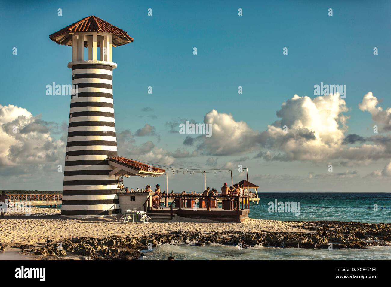 Dominicus, république dominicaine 20 août 2025 : touristes dégustant des cocktails dans un bar à côté d'un pittoresque phare rayé noir et blanc sur la beac Banque D'Images