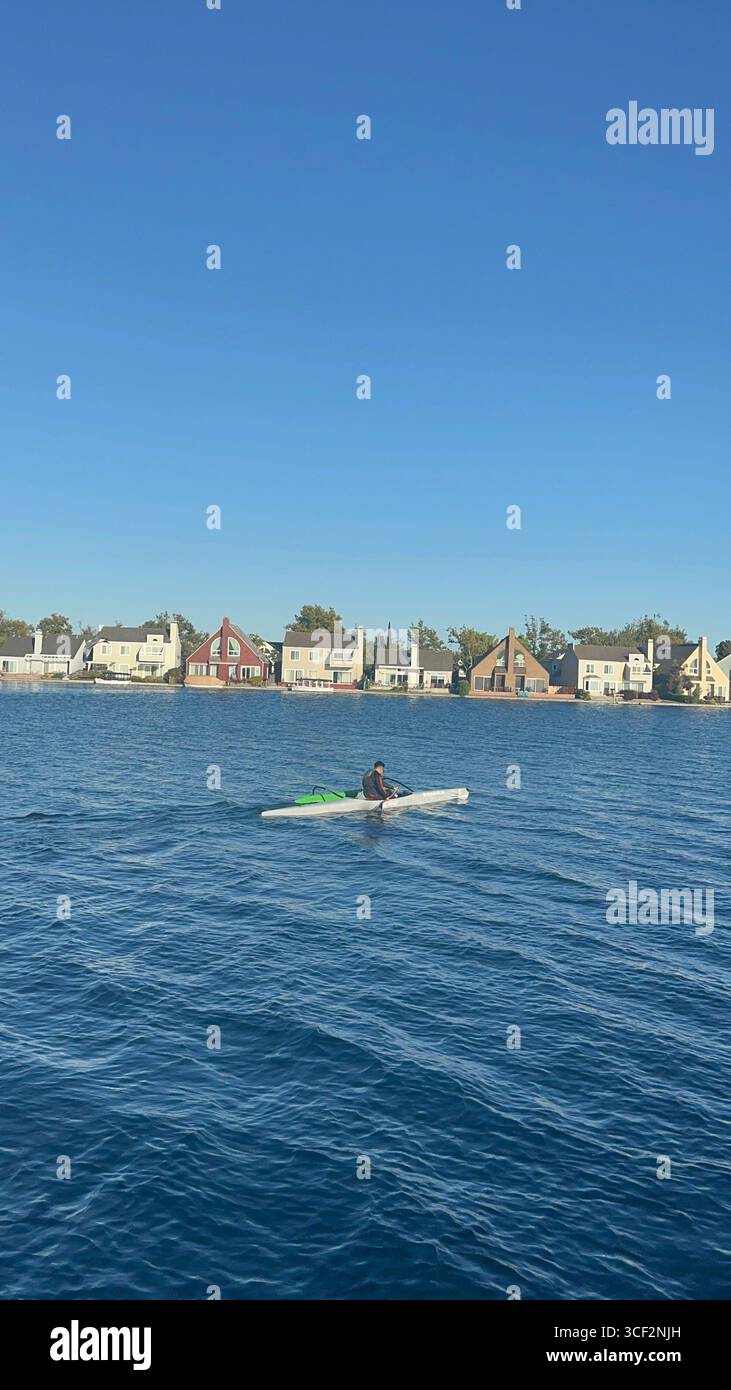 Un kayakiste pagaie sur l'eau bleue calme tandis qu'une mouette vole au-dessus de lui, avec des maisons au bord de l'eau et des immeubles de bureaux modernes en arrière-plan - Image de stock capturée avec un smartphone