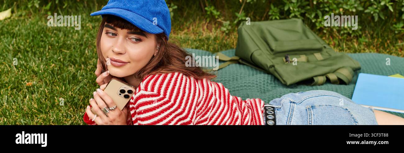 Une belle jeune femme est allongée sur un champ herbeux, étudiant et souriant tout en utilisant son téléphone. Banque D'Images