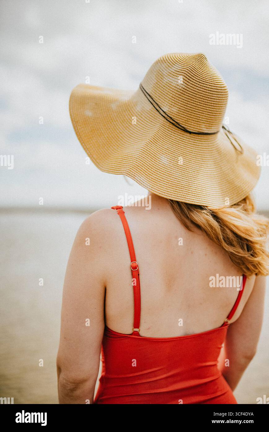 Une femme portant un haut rouge et un chapeau de paille se tient debout sur une plage. Le chapeau est grand et a un rebord jaune Banque D'Images