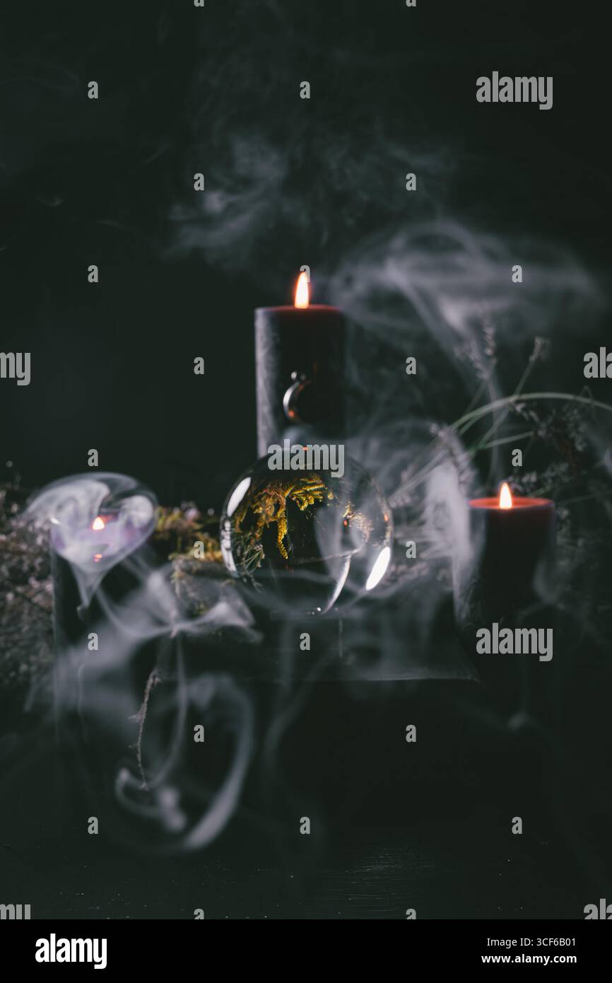 Boule de cristal entourée de bougies noires brûlantes et de fumée tourbillonnante, évoquant une atmosphère mystérieuse et magique dans un espace rituel sombre Banque D'Images