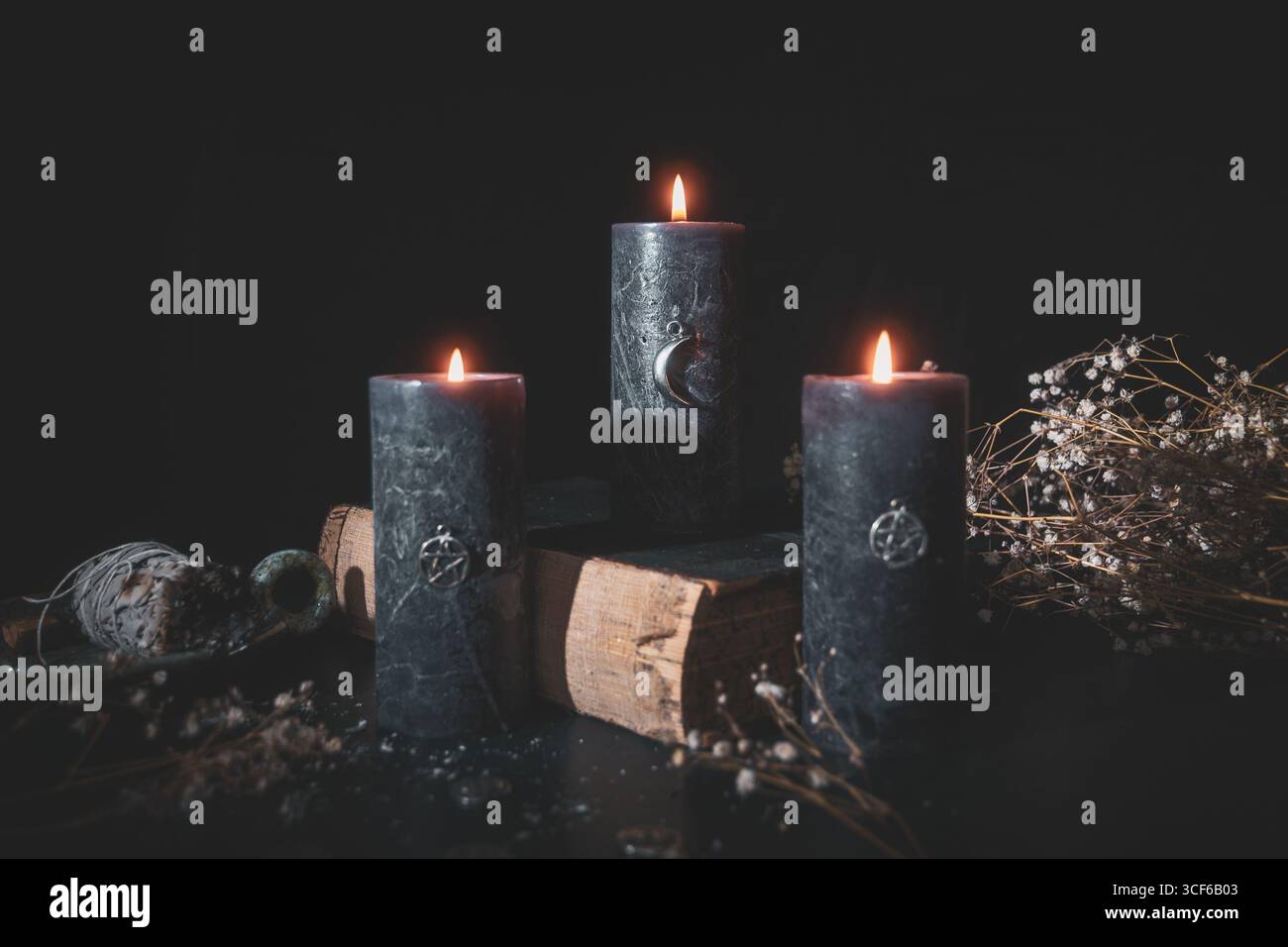 Trois bougies rituelles noires avec des charmes allumées à côté d'herbes et de vieux livres, créant un autel de sorcellerie mystique dans une ambiance sombre. Banque D'Images