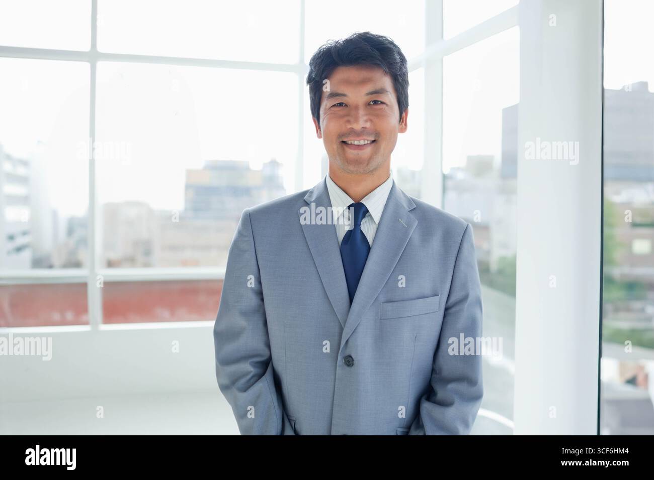 Homme japonais portant un costume gris et une cravate bleue debout dans un bureau lumineux avec des baies vitrées Banque D'Images