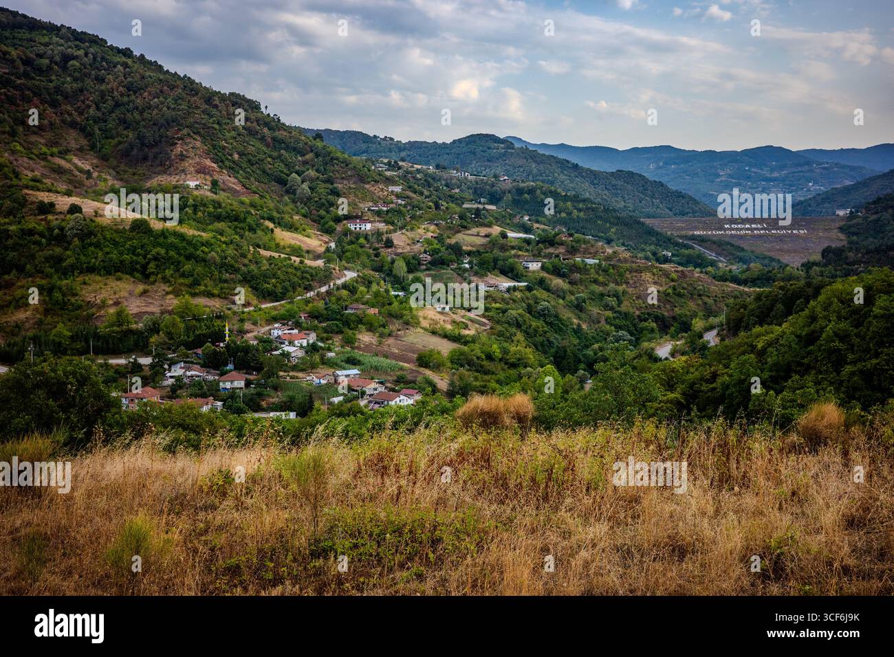 Vallée pittoresque à Başiskele, Kocaeli, Türkiye, avec des maisons de village dispersées, des collines verdoyantes et de l'herbe sèche sous un ciel partiellement nuageux. Banque D'Images