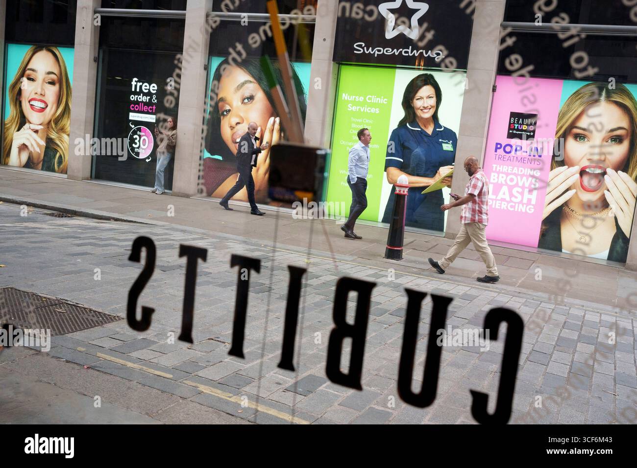 Les employés de la ville passent devant les visages de femmes tapissant l'extérieur d'une succursale récemment ouverte de la marque de pharmacie Superdrug, dans la ville de Londres, le quartier financier de la capitale, le 20 août 2025, à Londres, en Angleterre. Banque D'Images