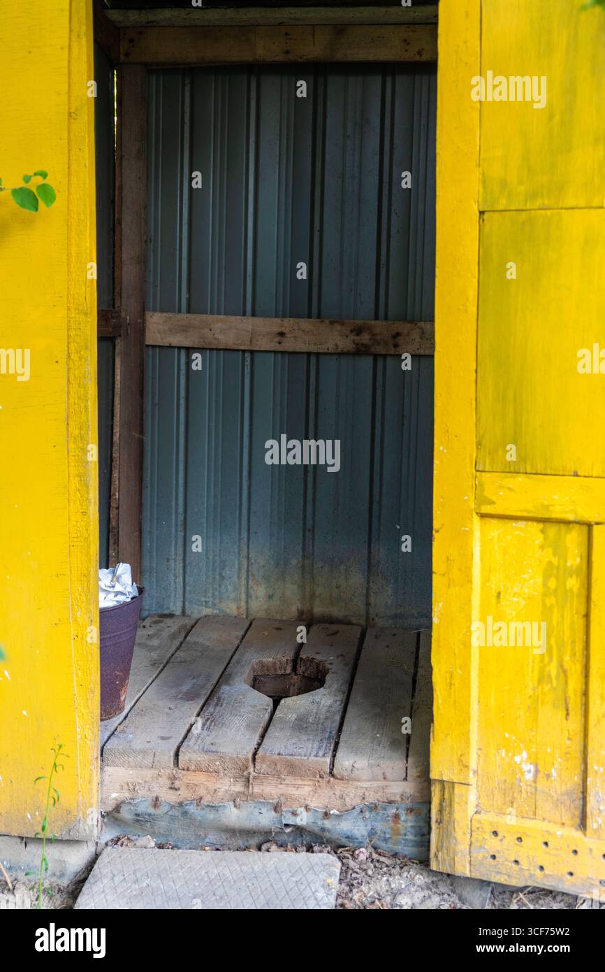 Terrasse ouverte en bois avec porte jaune et mur métallique, avec un simple trou de toilette, cadre rural, lumière naturelle. Banque D'Images