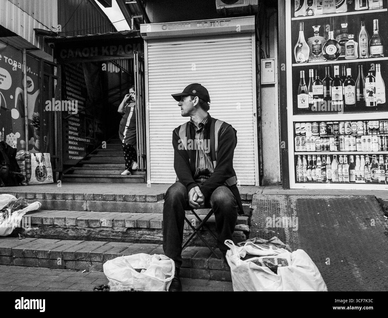 Kiev, Ukraine - 20 juillet 2025 : vendeur de rue assis sur une chaise pliante devant son stalle avec des sacs de marchandises à ses pieds, tandis qu'une femme monte les escaliers dans un marché aux puces ukrainien Banque D'Images