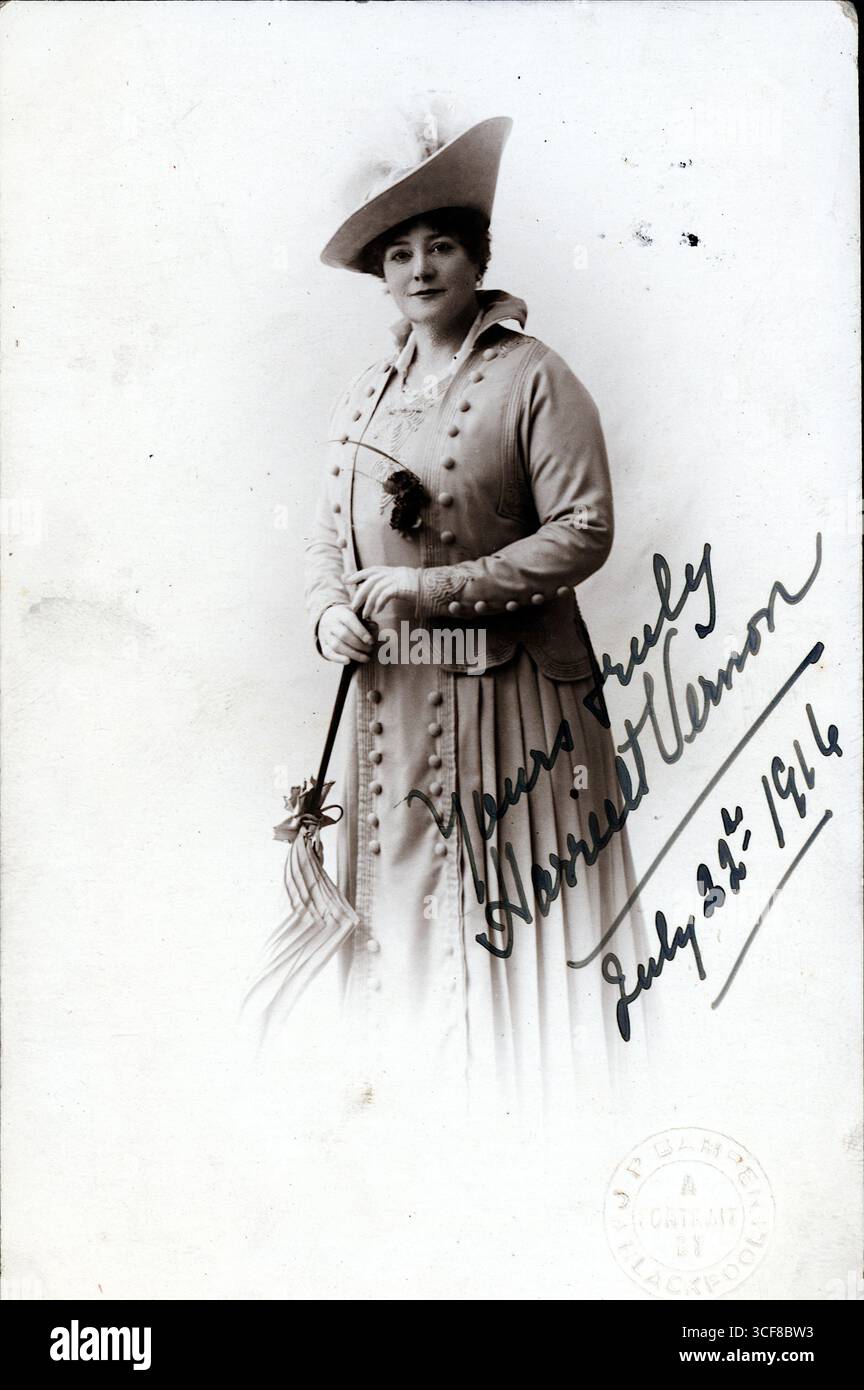 Harriet Vernon, célèbre interprète de Music-hall, se tient dans une élégance serrée, son parasol incliné avec grâce et son chapeau à plumes prêtant un flair dramatique. La robe boutonnée et la jupe plissée reflètent la mode des années 1910, tandis que sa signature manuscrite – datée du 25 juillet 1916 – ancre l’image dans la Grande-Bretagne en temps de guerre. Photographié par Bamber de Church Street, Blackpool, le portrait mêle célébrité et décorum, capturant le charisme de Vernon dans un moment de calme dignité théâtrale. Le timbre studio circulaire affirme sa provenance comme un souvenir régional de résonance culturelle. Banque D'Images