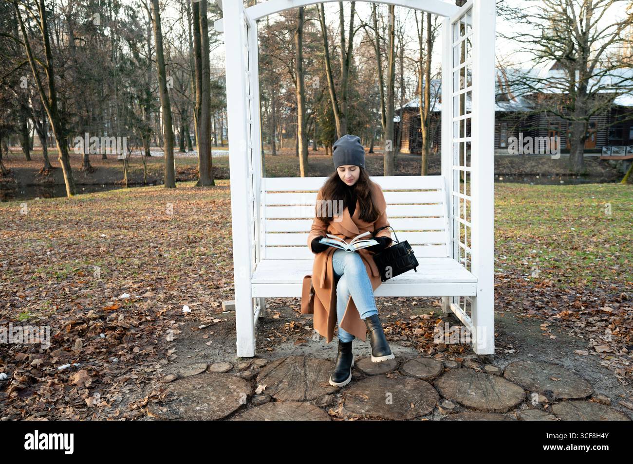 Lecture sous les arbres dans un parc serein sur une journée d'automne fraîche tout en étant confortable et élégant dans un manteau chaud et chapeau Banque D'Images