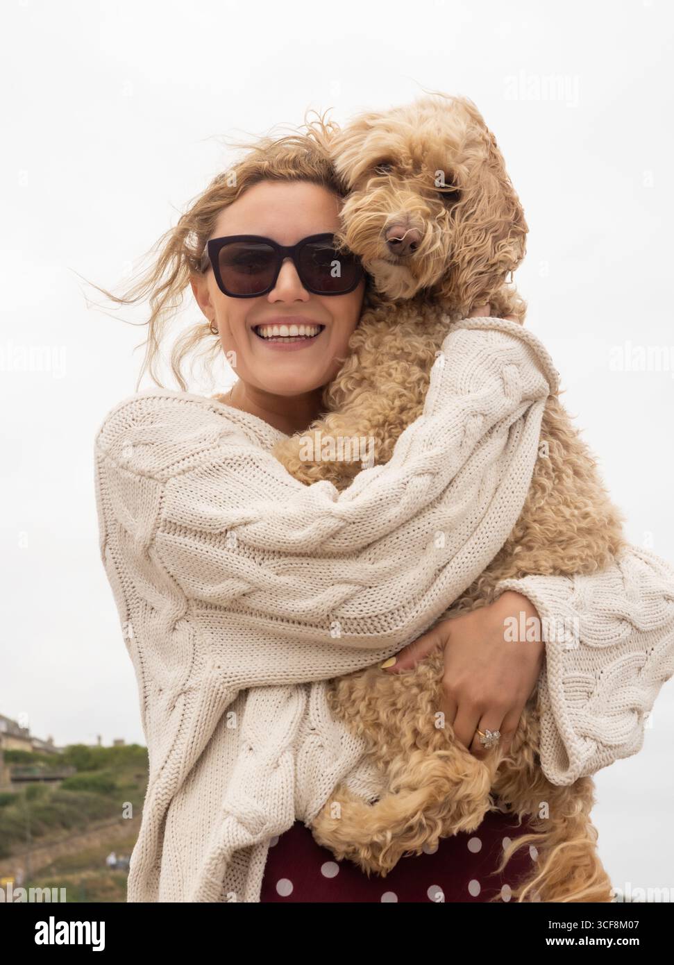 Une femme souriante portant un pull crème et des lunettes de soleil câlin son chien Cockapoo sur la plage de Saltburn-by-the-Sea, North Yorkshire, Royaume-Uni. Banque D'Images