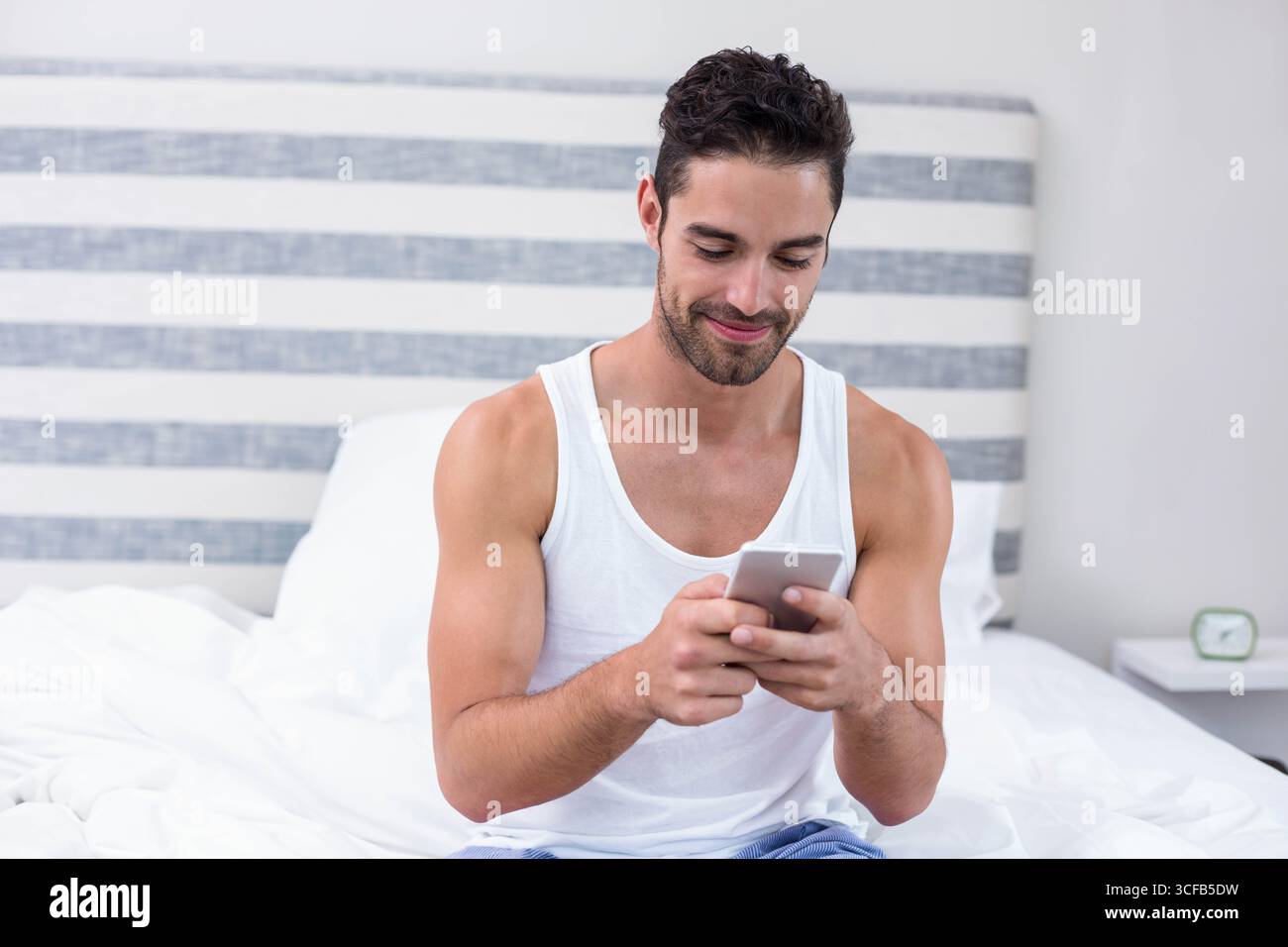 Homme vérifiant smartphone tout en étant assis sur le lit dans la chambre avec table de chevet et réveil analogique Banque D'Images