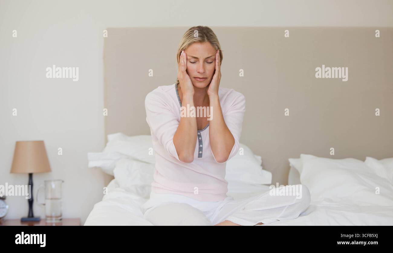 Femme assise sur le lit dans la chambre portant un pyjama pressant les mains aux tempes par réveil Banque D'Images