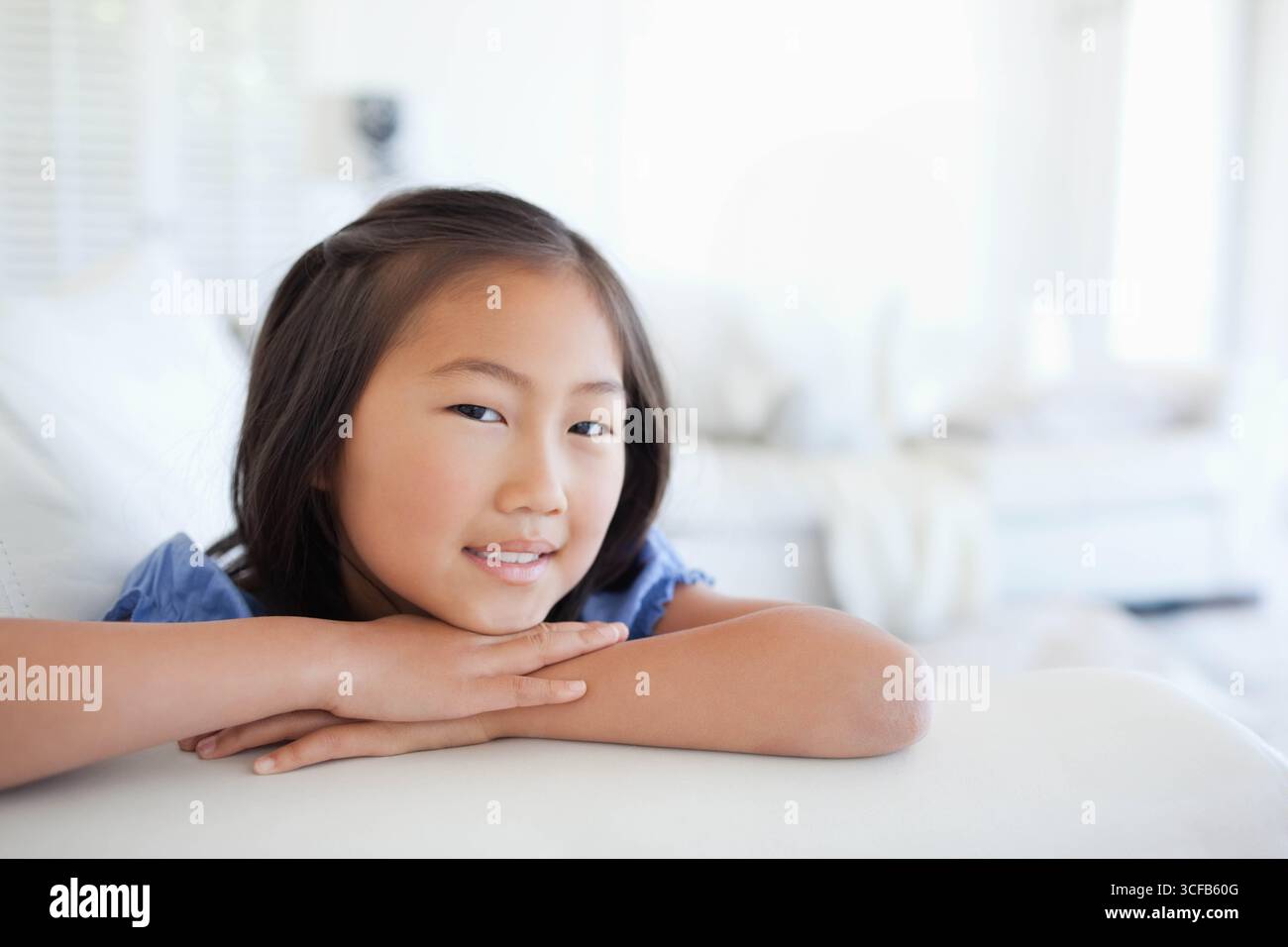 Fille asiatique d'âge scolaire s'inclinant sur un canapé blanc par des stores de fenêtre dans un salon lumineux, menton de repos Banque D'Images