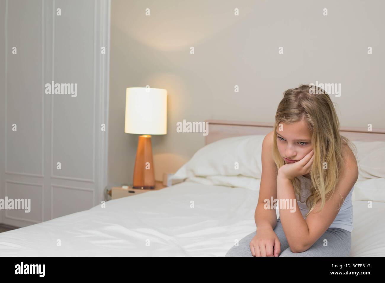 Fille enfant assise sur le bord du lit dans la chambre avec tête de lit en bois et lampe éclairée, espace copie Banque D'Images