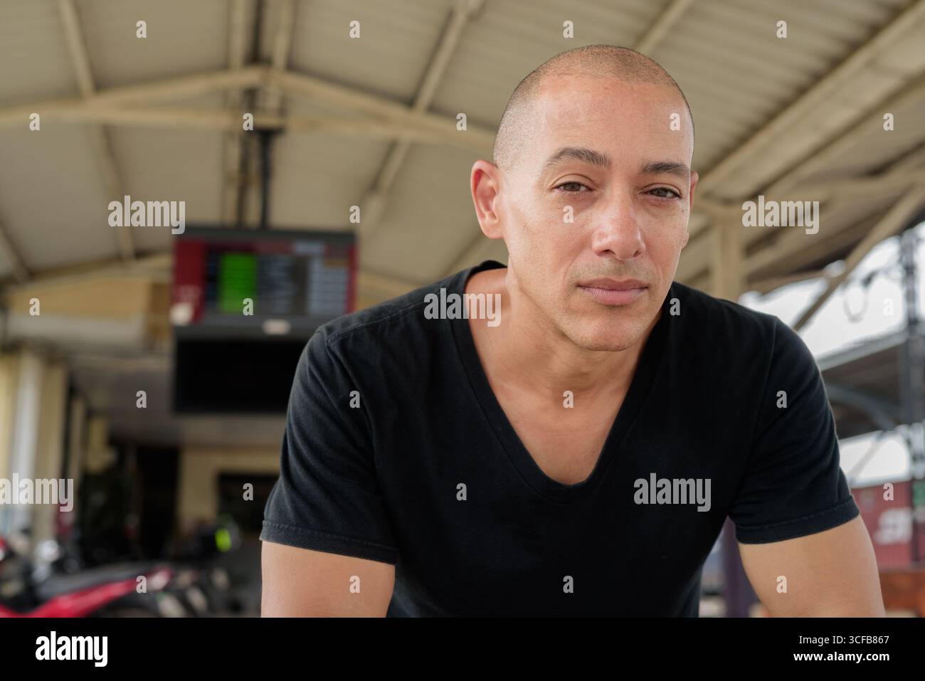 Portrait d'un touriste homme chauve hispanique mature dans un t-shirt noir assis à une plate-forme de gare, attendant un train lors d'un voyage en Thaïlande Banque D'Images