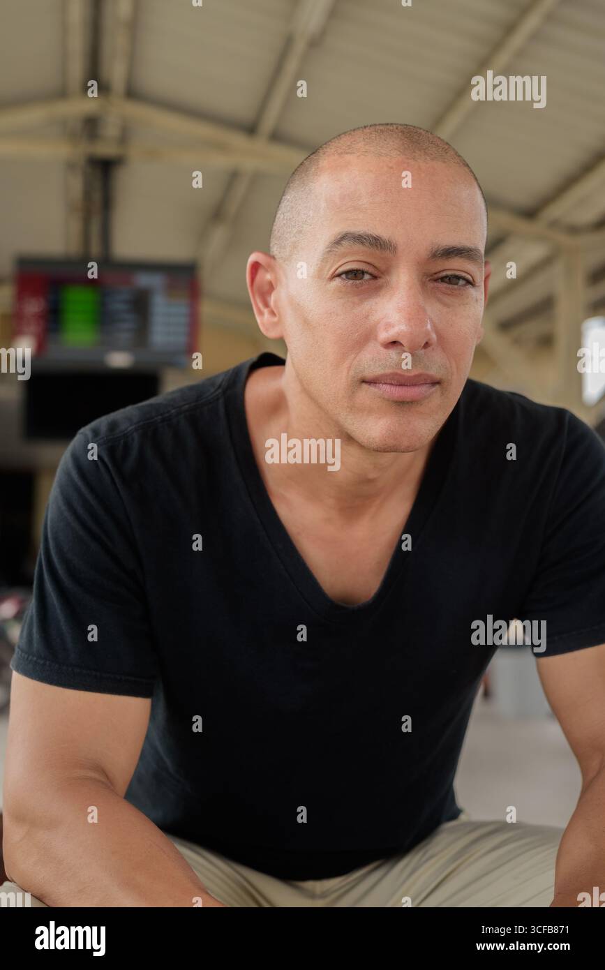 Portrait d'un touriste homme chauve hispanique mature dans un t-shirt noir assis à une plate-forme de gare, attendant un train lors d'un voyage en Thaïlande Banque D'Images