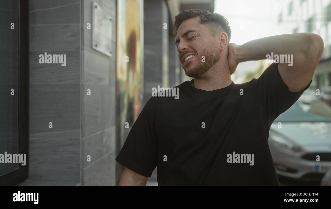 Jeune homme avec la barbe à l'extérieur dans la ville tenant le cou dans l'inconfort contre le fond urbain sous la lumière du soleil, portant chemise noire décontractée dans la rue pendant Banque D'Images