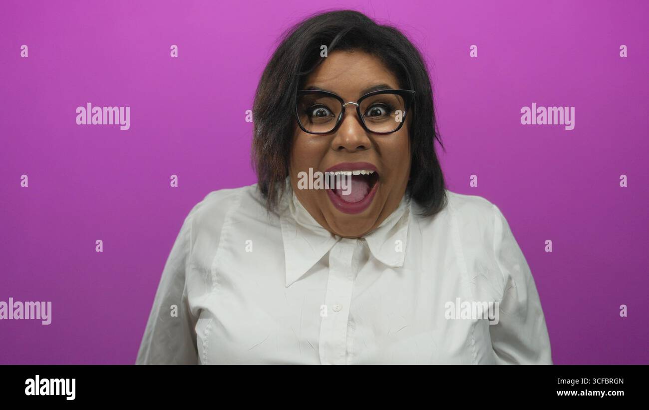 Femme avec des lunettes dans une chemise blanche exprimant la surprise sur un fond rose vif, soulignant ses émotions joyeuses et sa beauté grande taille. Banque D'Images
