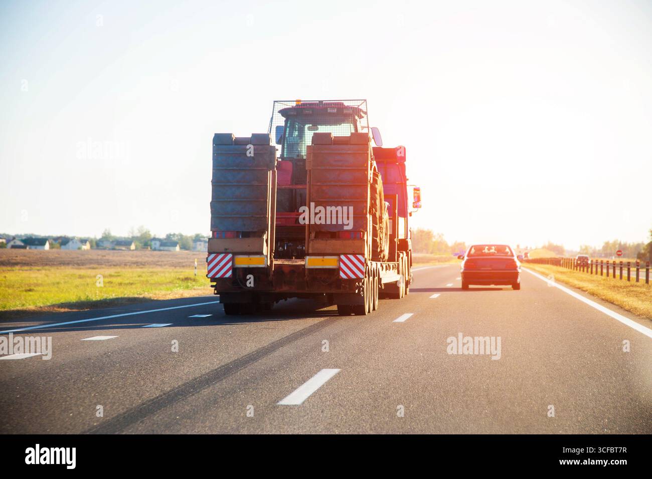 Transport d'un tracteur par un camion avec une semi-remorque lowboy sur une autoroute avec pour toile de fond un coucher de soleil en été. Transport de surdimensionnés Banque D'Images