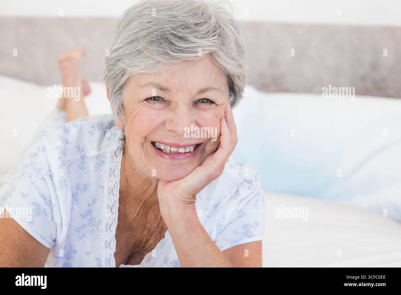 Femme aînée allongée sur le lit portant un dessus de sommeil floral dans la chambre avec des oreillers blancs et de la literie Banque D'Images