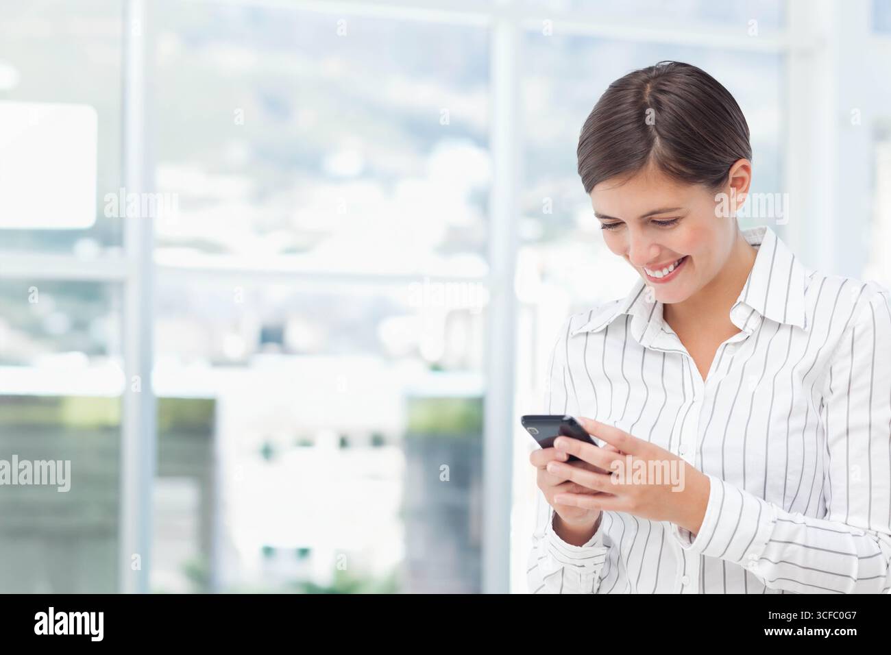 Employée de bureau portant une chemise rayée debout près de la fenêtre du hall pour vérifier l'espace de copie du smartphone Banque D'Images