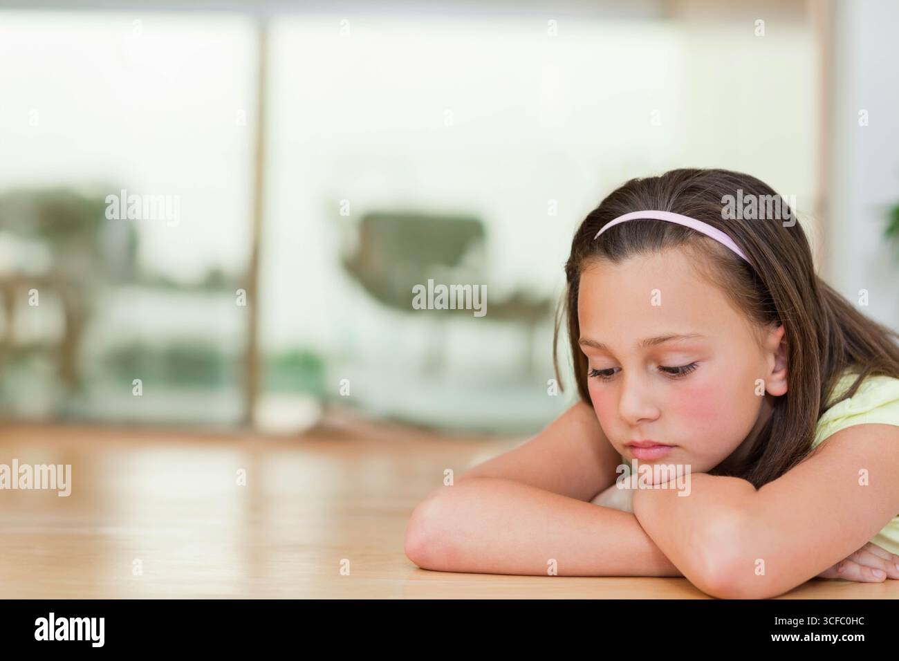 Fille d'âge scolaire se reposant sur les bras sur le plancher de bois près des portes vitrées portant un bandeau rose, espace copie Banque D'Images